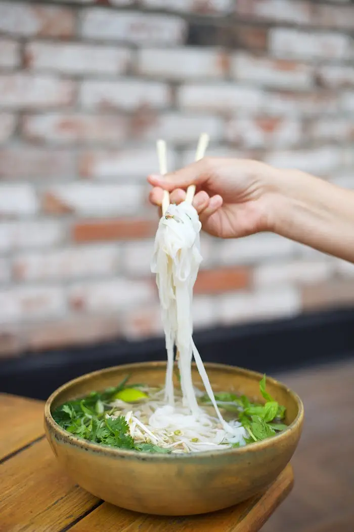 Hand holding chopsticks lifting noodles from a ceramic bowl of Asian soup with herbs.